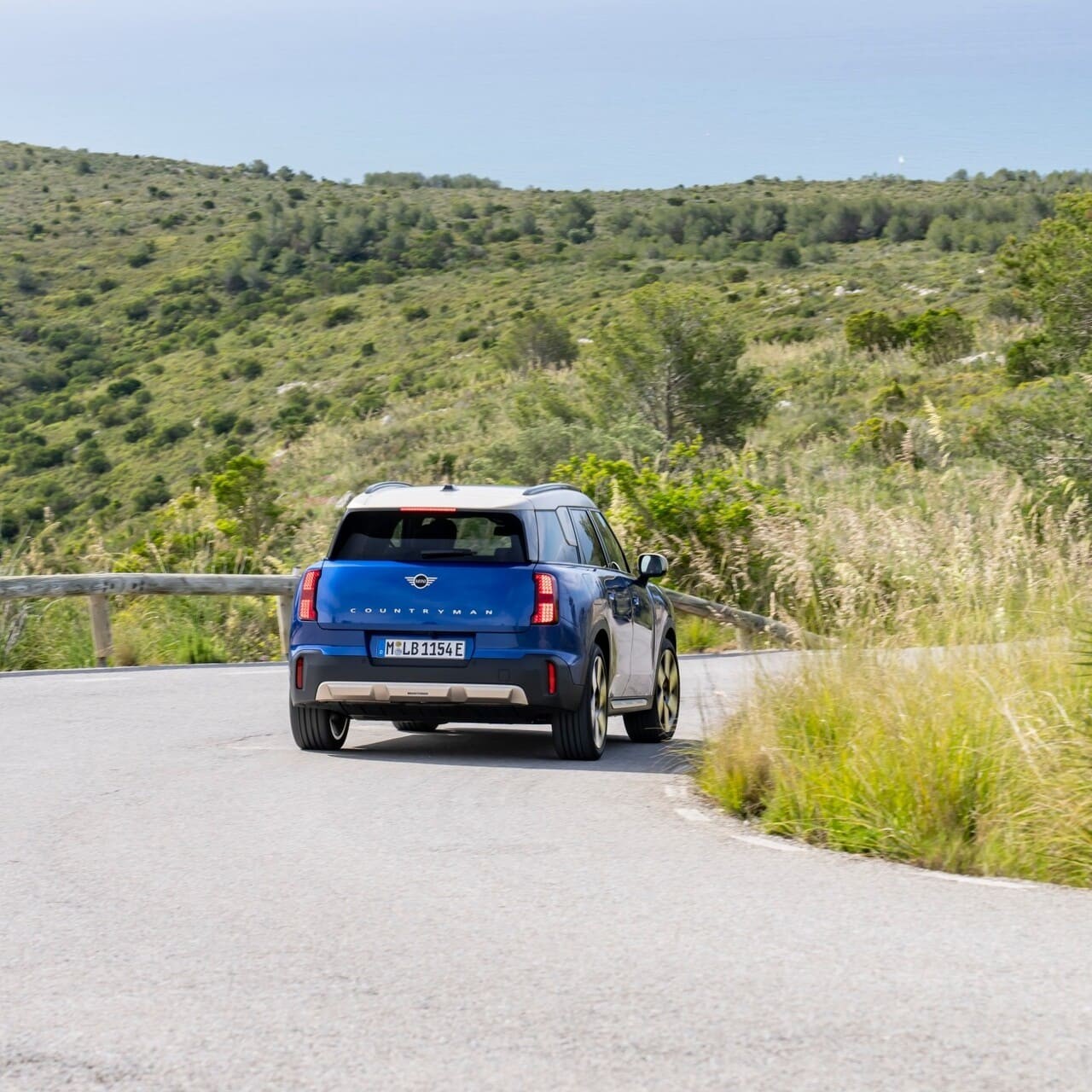 Vista traseira do MINI Countryman E a circular na estrada no meio de paisagem natural.